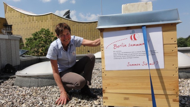 Eine Frau sitzt hockend vor einem urbanen Zuhause für Bienen, das aussieht wie ein kleines Holzhaus. Im Hintergrund sieht man die Berliner Philharmonie.